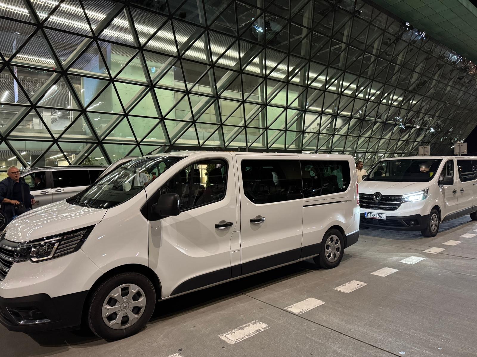 Flota białych busów Renault Trafic na tle nowoczesnego budynku z geometryczną fasadą, gotowa do przewozu osób.