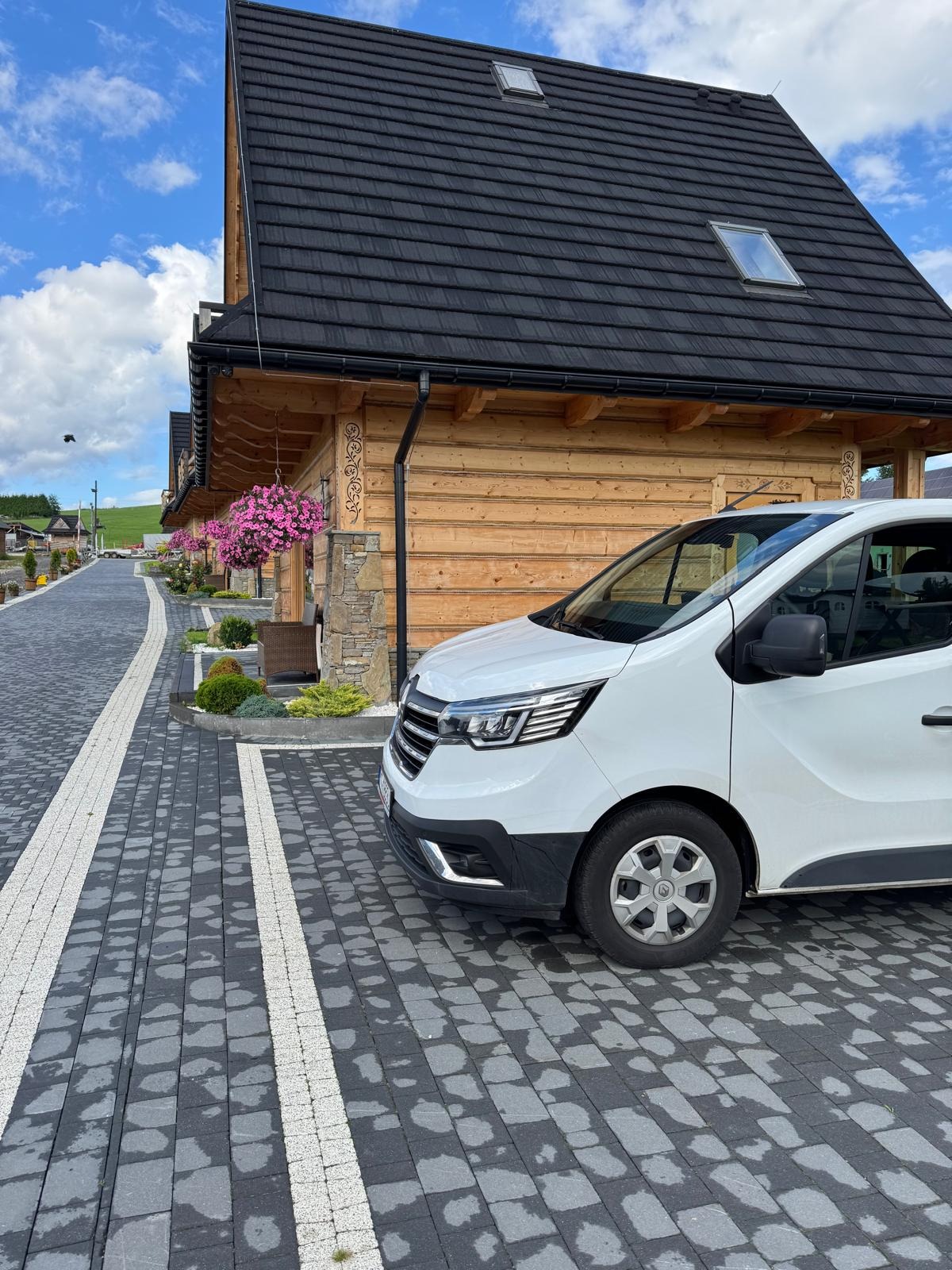 Biały van na parkingu z kostki brukowej przed drewnianym domem w stylu góralskim z kwiatami. Perspektywa z boku, widoczne detale domu i otoczenie.