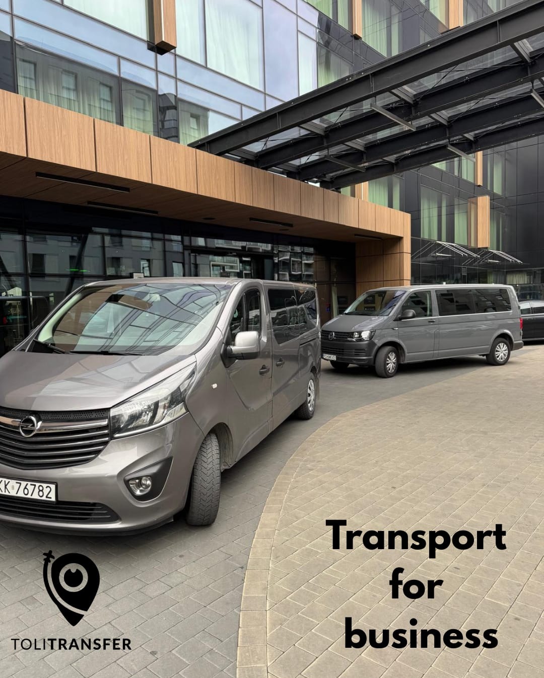 Dwa szare busy przed nowoczesnym budynkiem z przeszkloną fasadą i drewnianymi elementami. Jeden bus częściowo widoczny z przodu, drugi w tle.