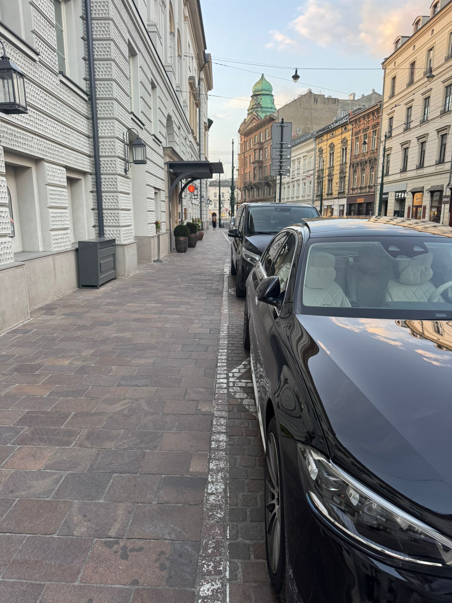 Dwa czarne samochody zaparkowane przy chodniku z kostki brukowej w centrum miasta. W tle budynki z zieloną kopułą i ozdobnymi fasadami.