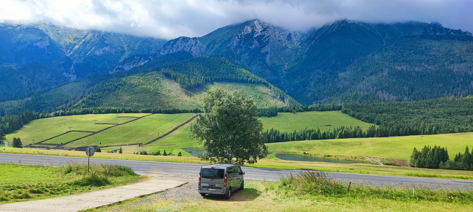 Szary van zaparkowany przy drodze z widokiem na zielone wzgórza i góry skryte pod chmurami, sugerujący podróż lub wycieczkę.