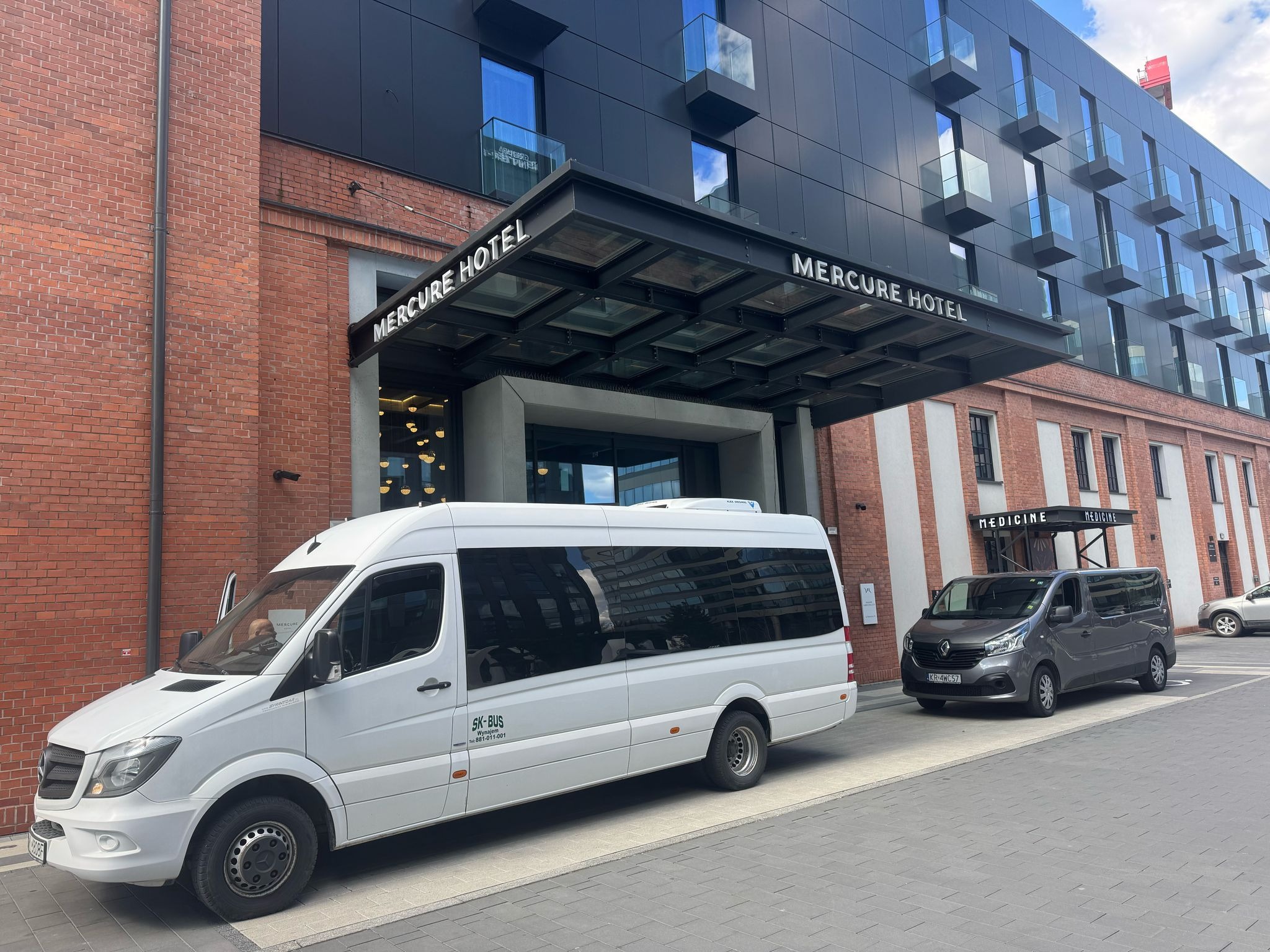 Dwa busy, biały i szary, zaparkowane przed hotelem Mercure. Biały bus z otwartymi drzwiami. Na drugim planie budynek z cegły i nowoczesna bryła hotelu.