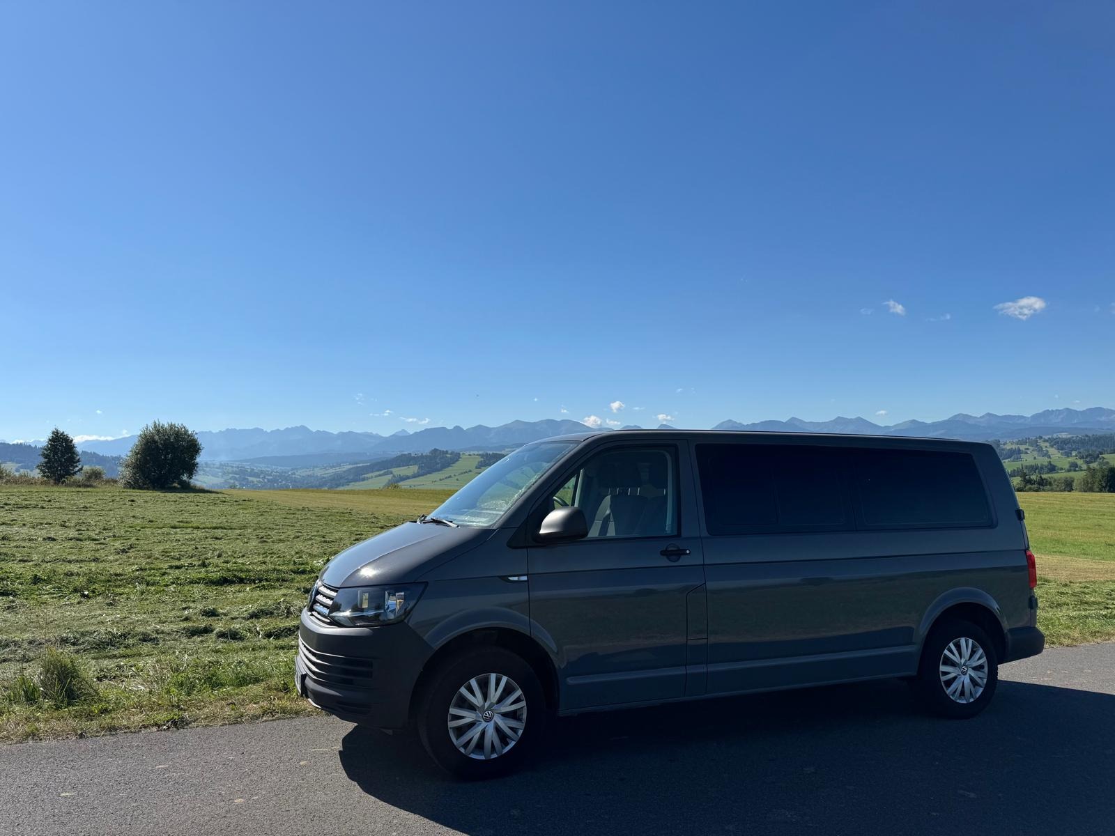 Szary bus VW Transporter na tle zielonych wzgórz i gór pod błękitnym niebem. Widok z boku, na pierwszym planie asfaltowa droga.