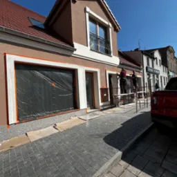 Dwóch pracowników na rusztowaniu podczas malowania elewacji budynku, okna zabezpieczone folią, widoczny fragment czerwonego pickupa Ford Ranger z polską tablicą rejestracyjną