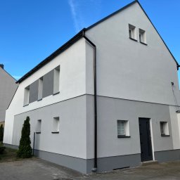 Fil-Bud - Dwukolorowa elewacja domu w odcieniach szarości i bieli, z czarnymi rynnami i drzwiami wejściowymi. Nowoczesny design, schludne wykończenie, błękitne niebo w tle.