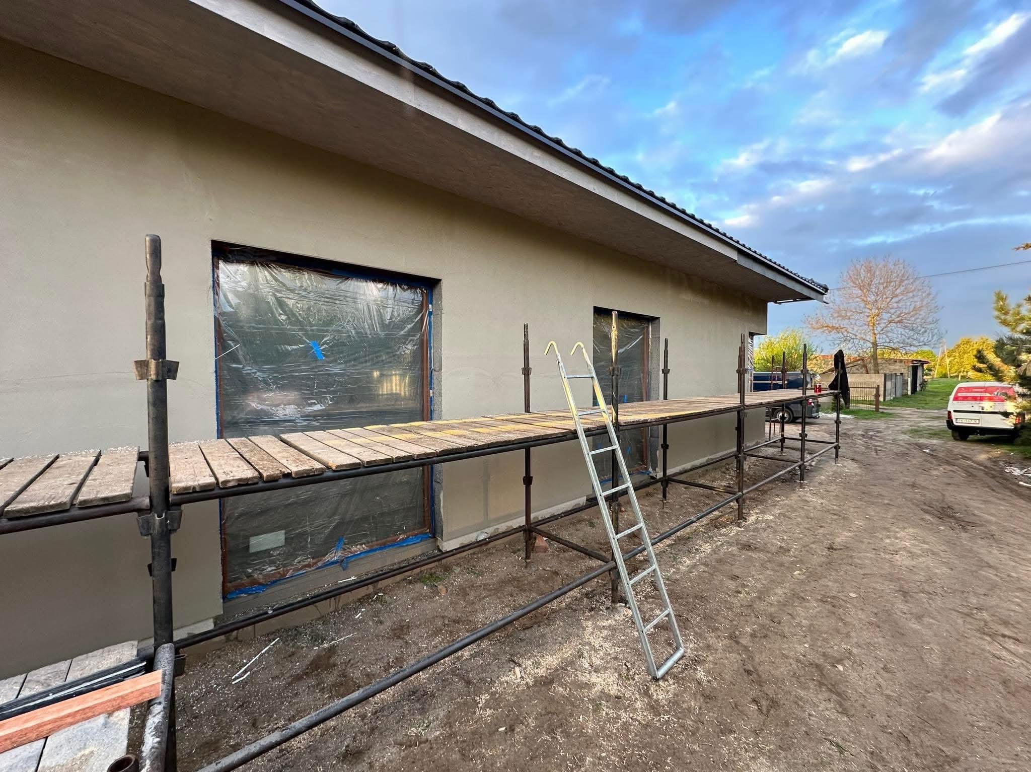 Świeżo otynkowana elewacja domu jednorodzinnego, zabezpieczone okna folią, widoczne rusztowanie z drewnianym pomostem i drabina, przygotowania do dalszych prac.