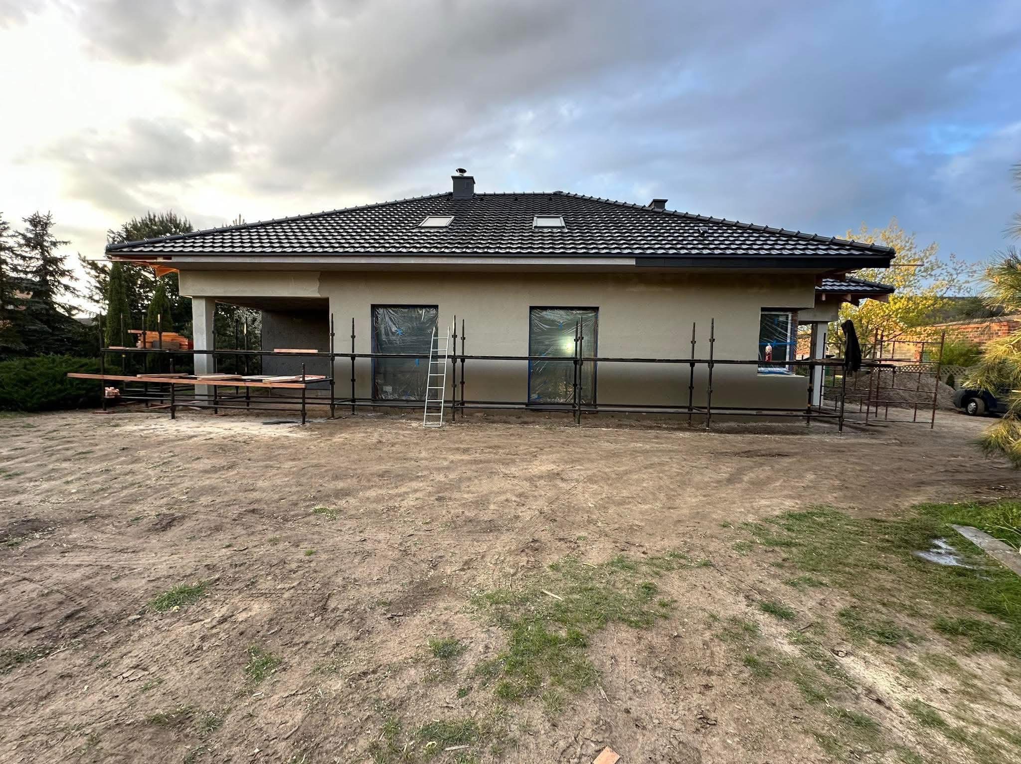 Dom jednorodzinny w trakcie ocieplania elewacji, widoczne rusztowania, okna zabezpieczone folią, częściowo otynkowana ściana, pochmurne niebo.