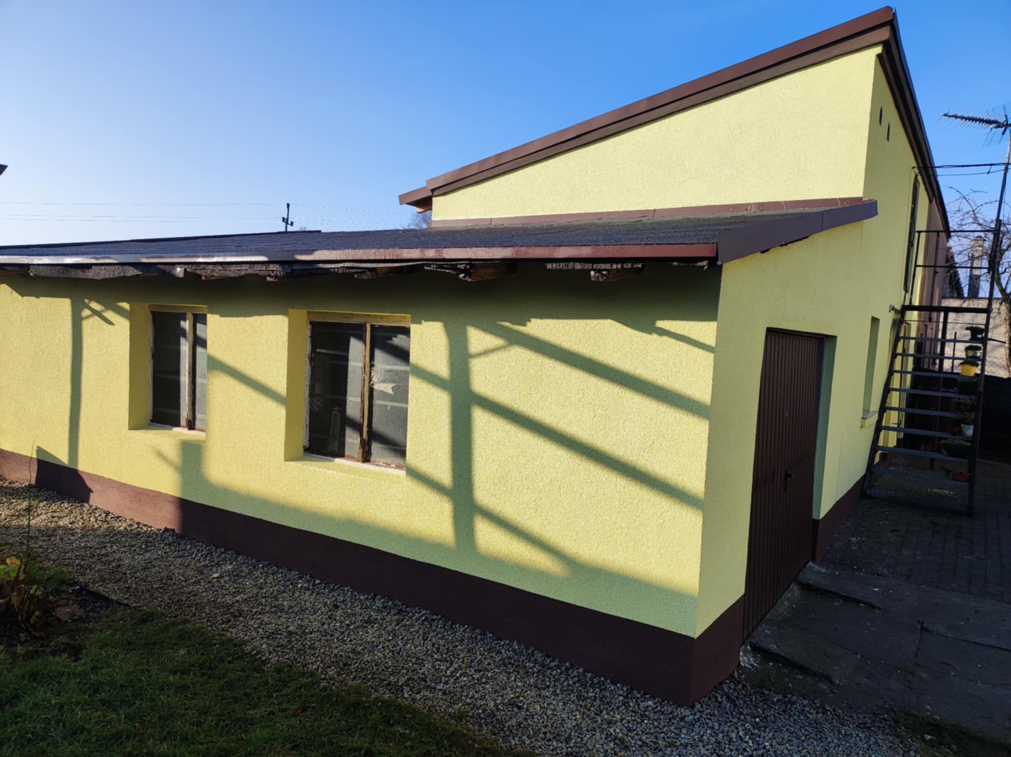 Żółta elewacja budynku gospodarczego z brązowymi detalami, widoczne cienie drzew, częściowo zniszczony dach i zewnętrzna klatka schodowa.