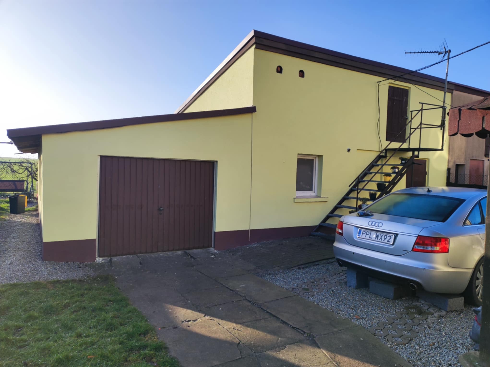 Żółty budynek gospodarczy z brązowymi akcentami, garażem i zewnętrznymi schodami, obok zaparkowany srebrny samochód Audi.