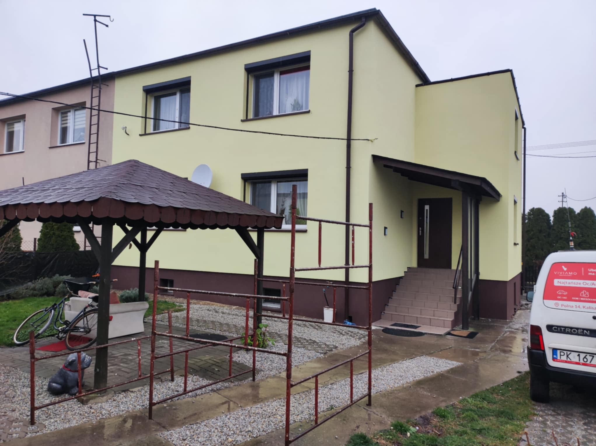 Dwupiętrowy dom z żółtą elewacją i brązowymi akcentami, widoczny z zewnątrz, z elementami dekoracyjnymi w ogrodzie i samochodem dostawczym firmy z reklamą na boku. W tle antena satelitarna.