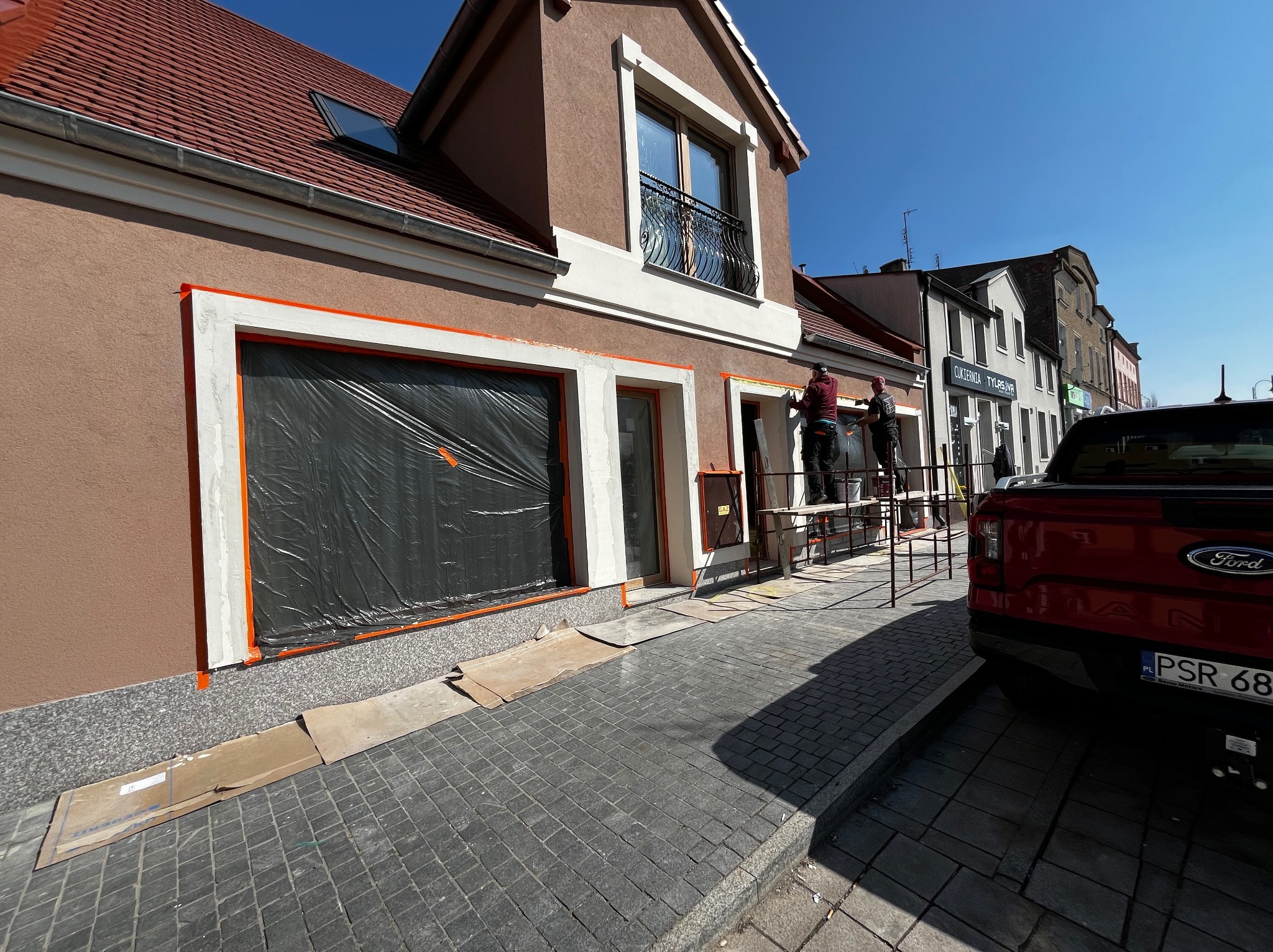 Dwóch pracowników na rusztowaniu podczas malowania elewacji budynku, okna zabezpieczone folią, widoczny fragment czerwonego pickupa Ford Ranger z polską tablicą rejestracyjną