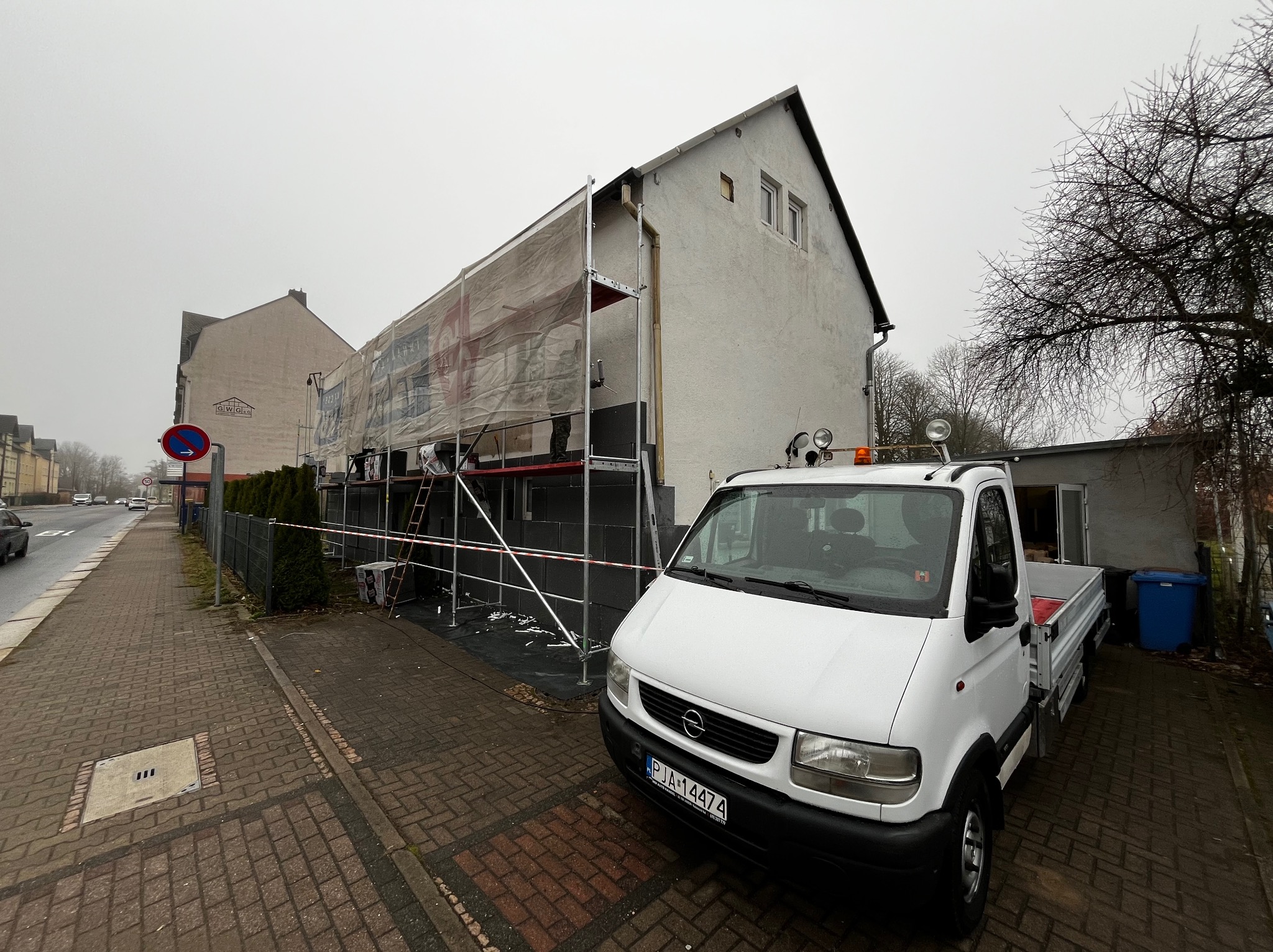 Dom jednorodzinny w trakcie ocieplania elewacji, widoczne rusztowania, zabezpieczająca siatka, płyty izolacyjne i zaparkowany biały samochód dostawczy z przodu.