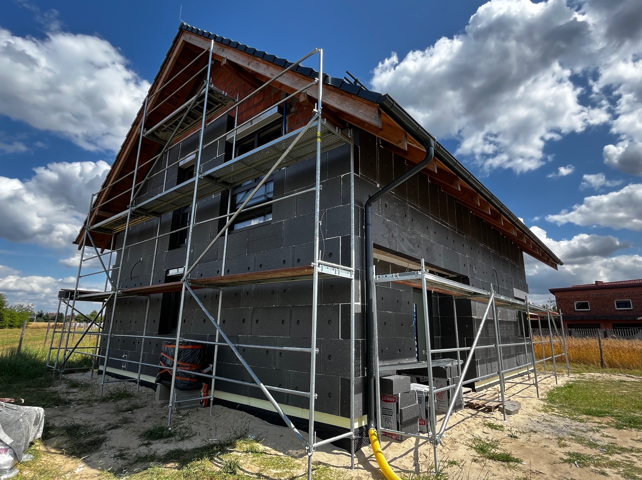 Dom w trakcie ocieplania, rusztowania na elewacji, widoczne czarne płyty izolacyjne. Niebieskie niebo z chmurami w tle. Dachówka na dachu.