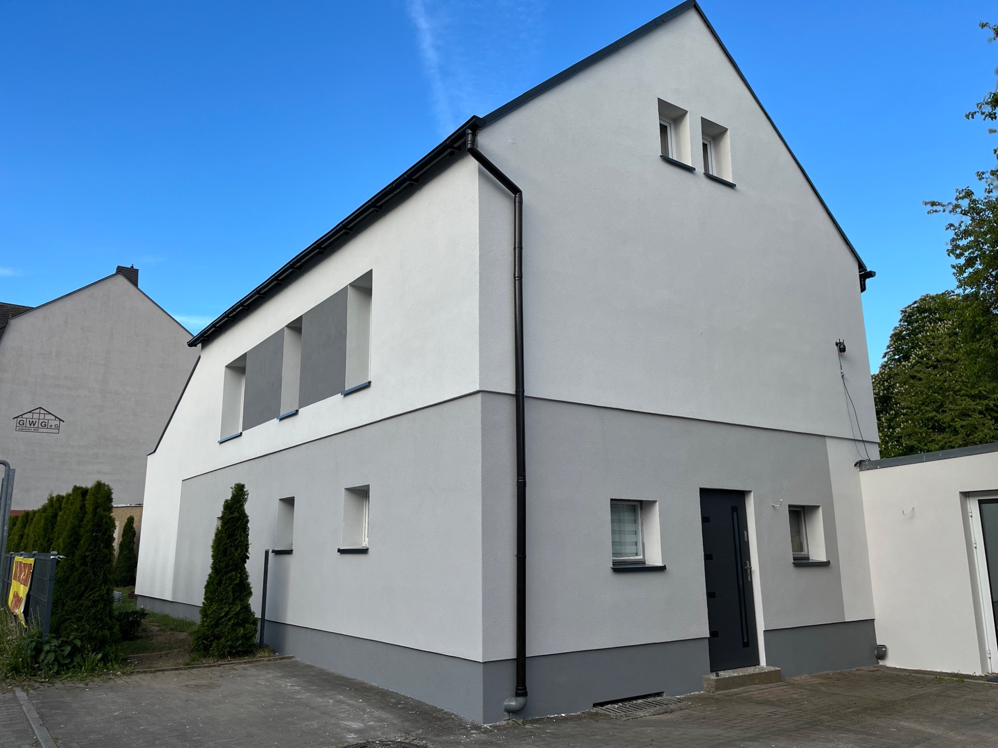 Dwukolorowa elewacja domu w odcieniach szarości i bieli, z czarnymi rynnami i drzwiami wejściowymi. Nowoczesny design, schludne wykończenie, błękitne niebo w tle.