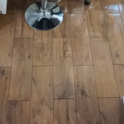 Ułożone panele podłogowe imitujące drewno, z widocznym fragmentem ściany z elementami dekoracyjnymi i chromowaną nogą krzesła barowego. Połyskująca powierzchnia.