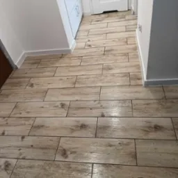 Ułożone podłużne płytki imitujące drewno na podłodze korytarza w Rudzie Śląskiej. Widoczne drzwi i białe listwy przypodłogowe. Płytki ułożone w poprzek korytarza.