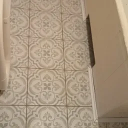 Ułożone płytki podłogowe w łazience, wzór w stylu retro, jasne kolory, widoczne białe listwy przypodłogowe i fragment toalety. Ujęcie z góry.