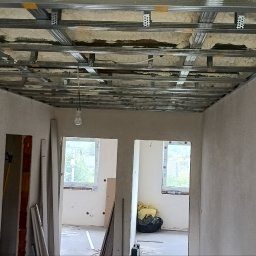 Rok - Sufit podwieszany z metalową konstrukcją i izolacją z wełny mineralnej, widok z dołu w trakcie prac wykończeniowych. W tle okna i surowe ściany.