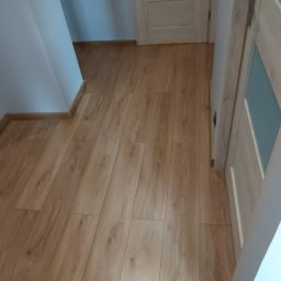 Rok - Jasne panele podłogowe ułożone w korytarzu, widoczne dwie pary drzwi z jasnego drewna. Ściany w odcieniach bieli i szarości. Widok z góry, ukośny kadr.