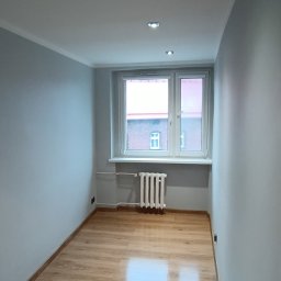 Rok - Wykończone wnętrze pokoju w Rudzie Śląskiej: panele podłogowe, pomalowane ściany na szaro, okno z widokiem na budynek. Minimalistyczny design, gotowe do zamieszkania.
