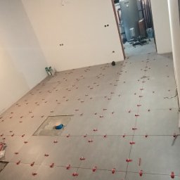 Rok - Szare płytki podłogowe w trakcie układania, z czerwonymi klipsami poziomującymi. Widoczny fragment instalacji podłogowej i surowy sufit w pomieszczeniu.