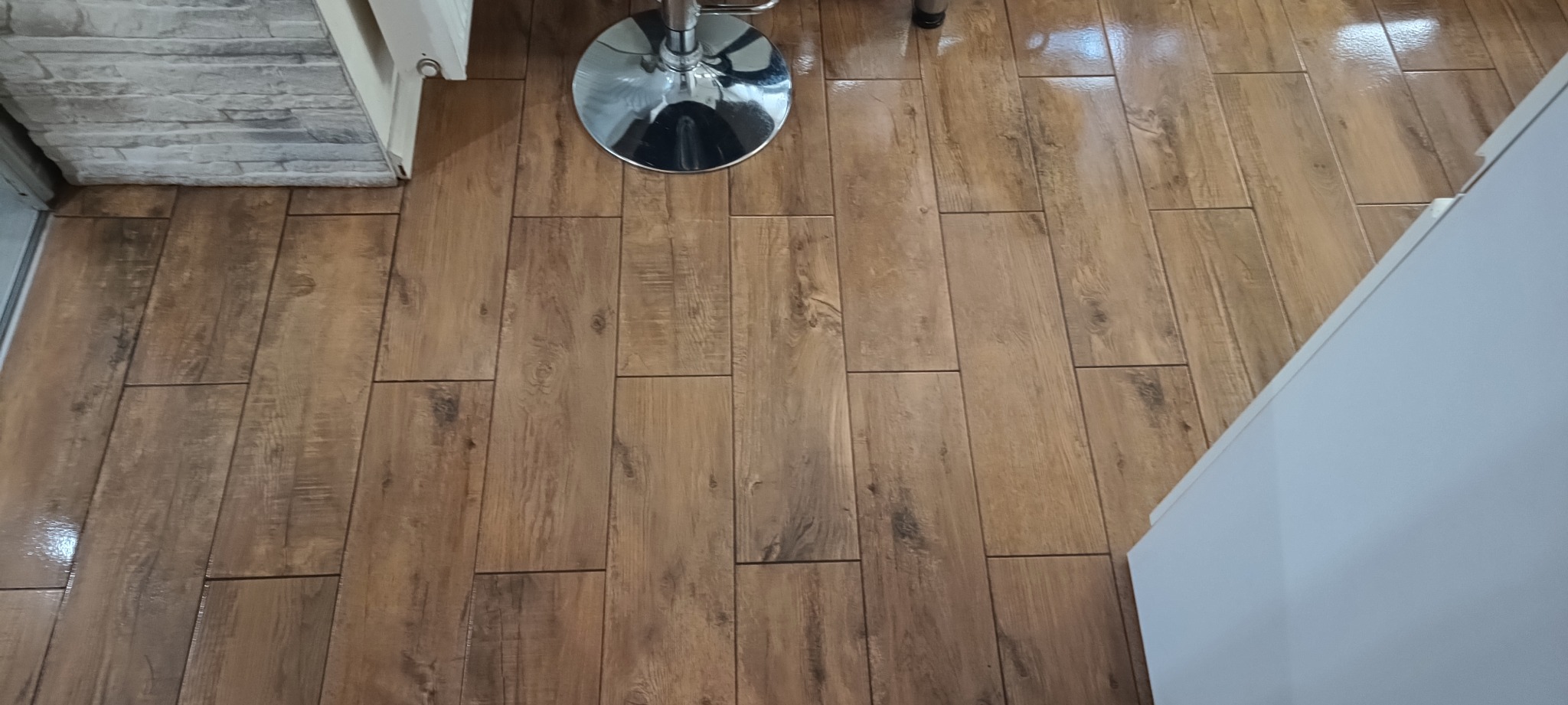 Ułożone panele podłogowe imitujące drewno, z widocznym fragmentem ściany z elementami dekoracyjnymi i chromowaną nogą krzesła barowego. Połyskująca powierzchnia.