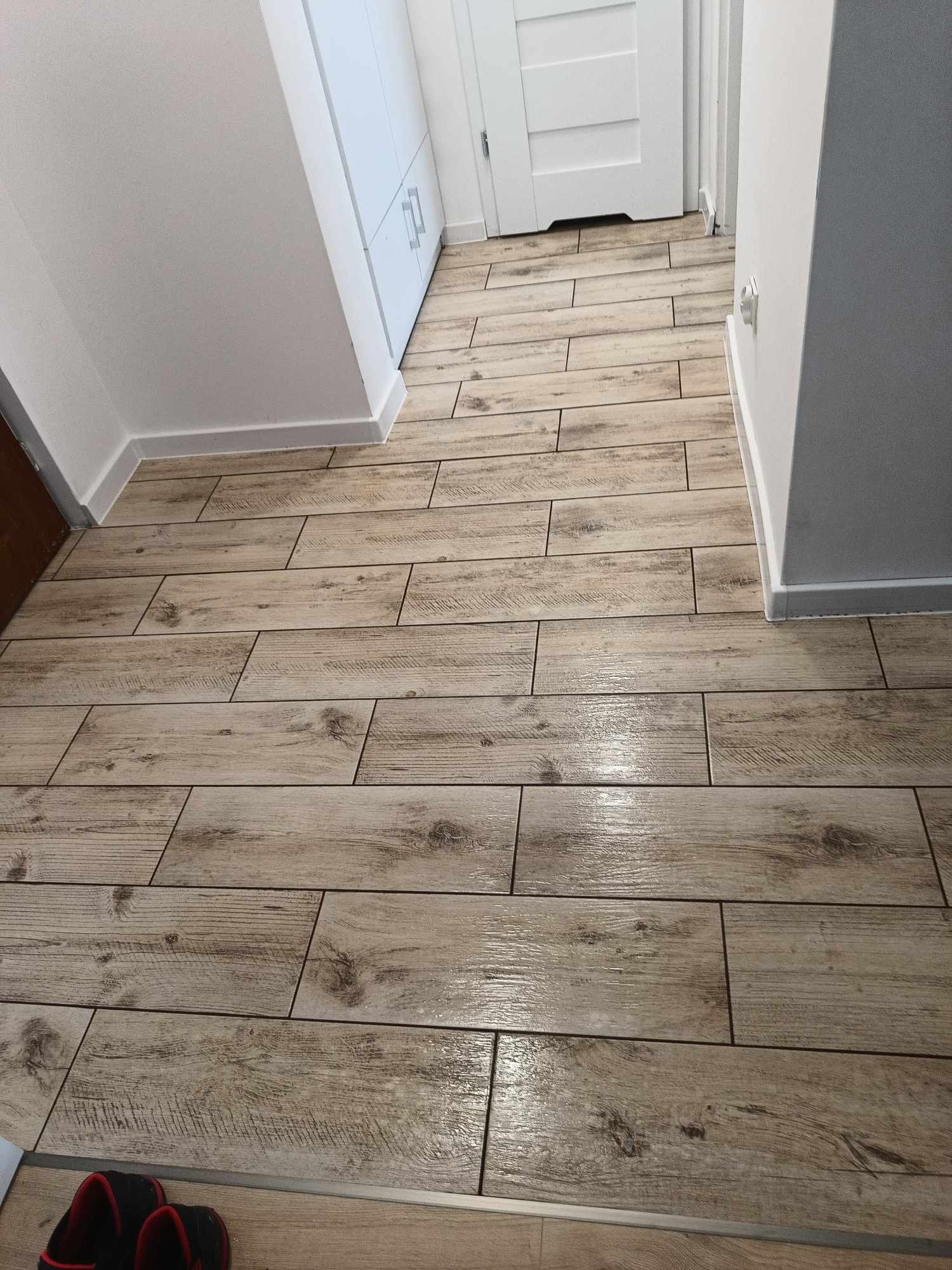 Ułożone podłużne płytki imitujące drewno na podłodze korytarza w Rudzie Śląskiej. Widoczne drzwi i białe listwy przypodłogowe. Płytki ułożone w poprzek korytarza.