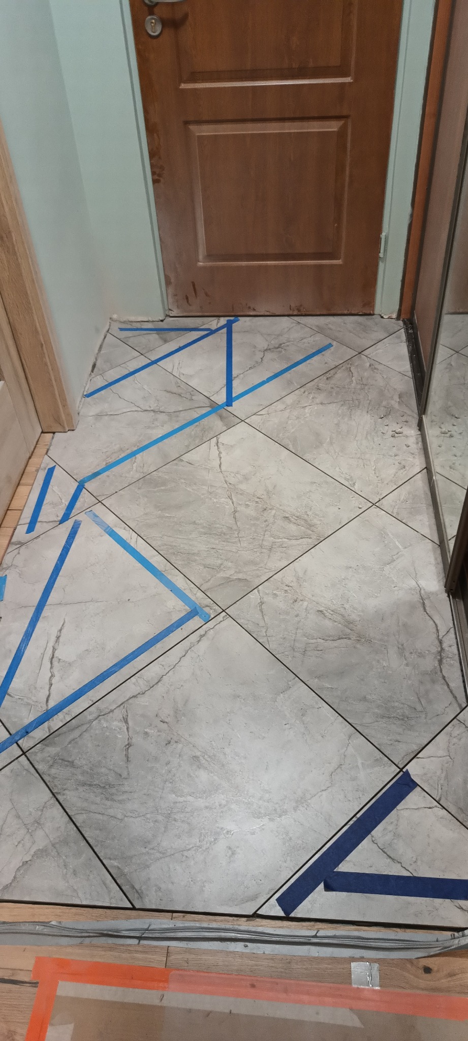 Ułożone szare płytki podłogowe z wzorem marmuru, zabezpieczone niebieską taśmą malarską, widok na próg drzwi wejściowych.