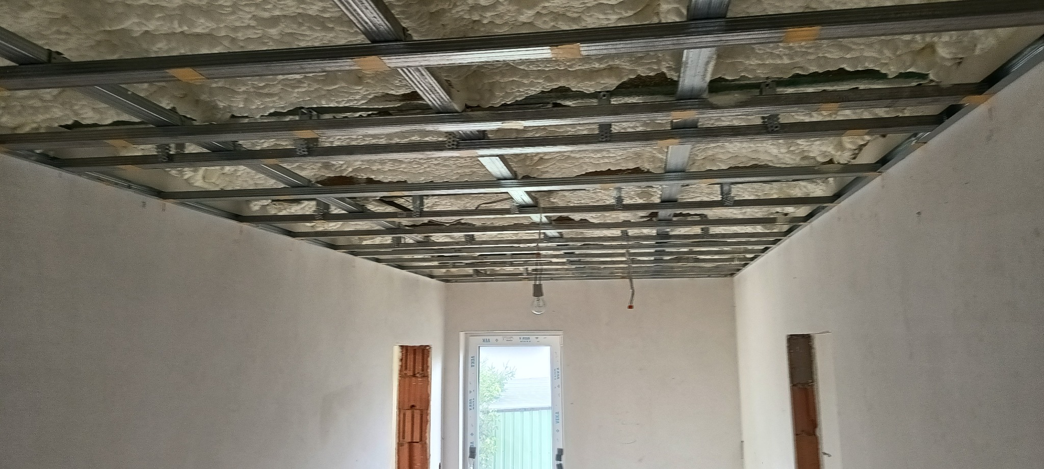 Montaż sufitu podwieszanego z płyt karton-gips: metalowa konstrukcja z izolacją z pianki, widoczne ściany w surowym stanie, okno. Etap prac wykończeniowych.