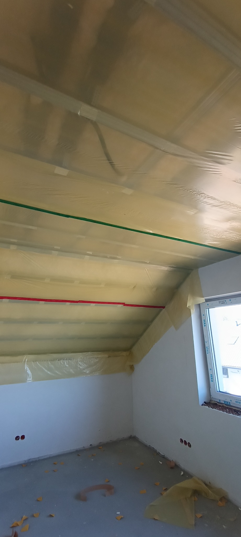 Pokój na poddaszu zabezpieczony folią malarską przed pracami wykończeniowymi. Widoczne profile sufitowe i instalacje. Okno z widokiem na zewnątrz.