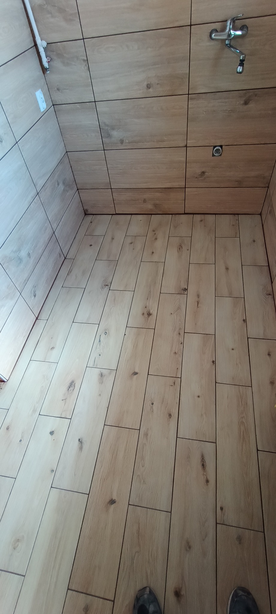 Wykończona łazienka z drewnopodobnymi płytkami na podłodze i ścianach. Widoczna bateria umywalkowa i fragment stopy osoby fotografującej. Jasne, ciepłe oświetlenie.