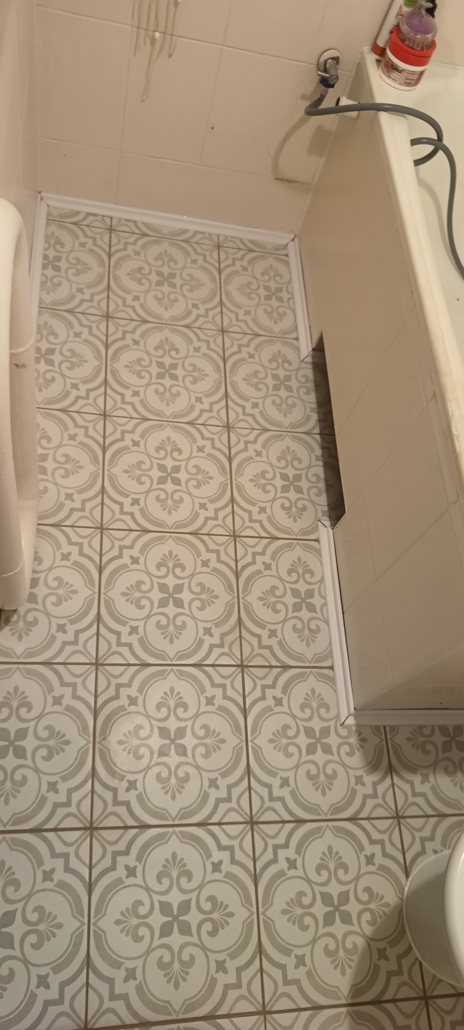 Ułożone płytki podłogowe w łazience, wzór w stylu retro, jasne kolory, widoczne białe listwy przypodłogowe i fragment toalety. Ujęcie z góry.