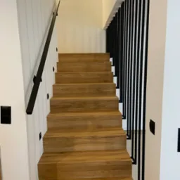 Drewniane schody w nowoczesnym wnętrzu z czarnymi, pionowymi balustradami i minimalistyczną poręczą. Oświetlenie punktowe na ścianie, jasne kolory ścian.