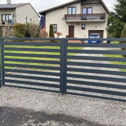Tul-Stal - Nowoczesna, pozioma brama wjazdowa z metalowych profili w kolorze grafitowym, zamontowana na posesji z domem i podjazdem wysypanym żwirem.