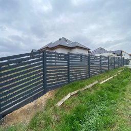Tul-Stal - Grafitowe ogrodzenie panelowe, poziome deski, świeżo po montażu, widok z zewnątrz posesji, trawa, domy w tle, pochmurne niebo.