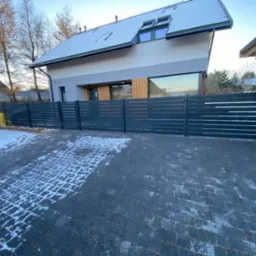 Nowoczesne, poziome ogrodzenie panelowe w kolorze antracytowym przed domem z szarym dachem, w zimowej scenerii. Widoczna kostka brukowa z resztkami śniegu.