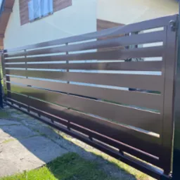 Nowoczesna, brązowa brama przesuwna z poziomymi panelami na tle domu z jasną elewacją i brązowym wykończeniem. Widoczne ogrodzenie panelowe po lewej stronie.