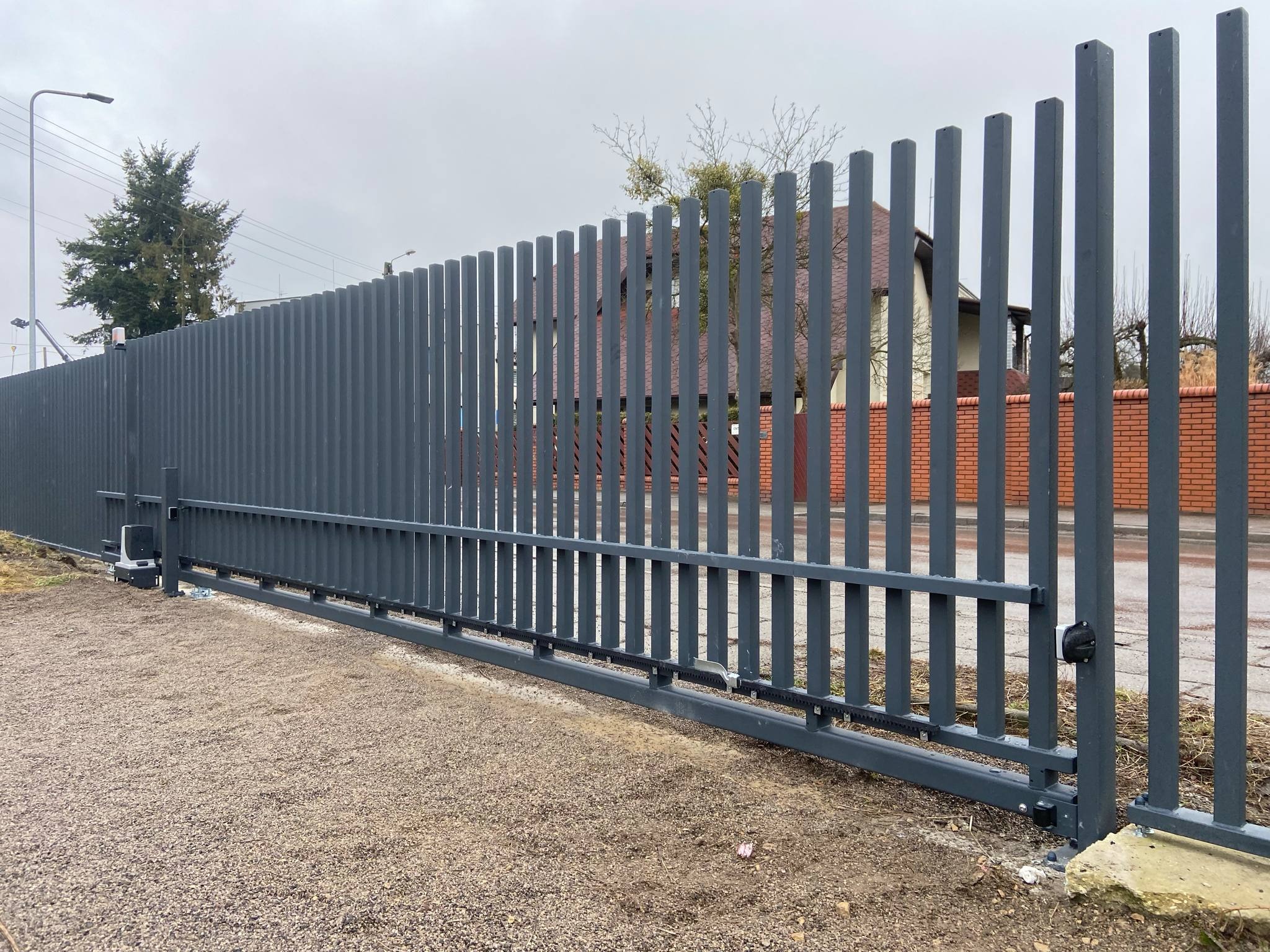 Nowoczesna, przesuwna brama wjazdowa z pionowych, grafitowych profili stalowych. Widoczny automat do bramy i fragment ogrodzenia z cegły w tle. Ujęcie z poziomu gruntu.