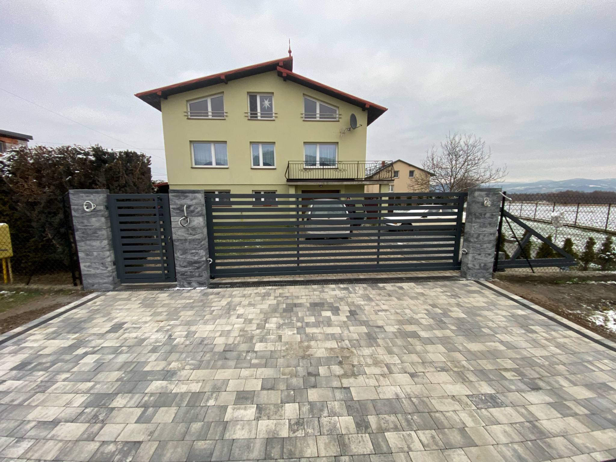 Nowoczesna brama wjazdowa z poziomych profili, szara kostka brukowa na podjeździe, dwukondygnacyjny dom w tle, pochmurne niebo.