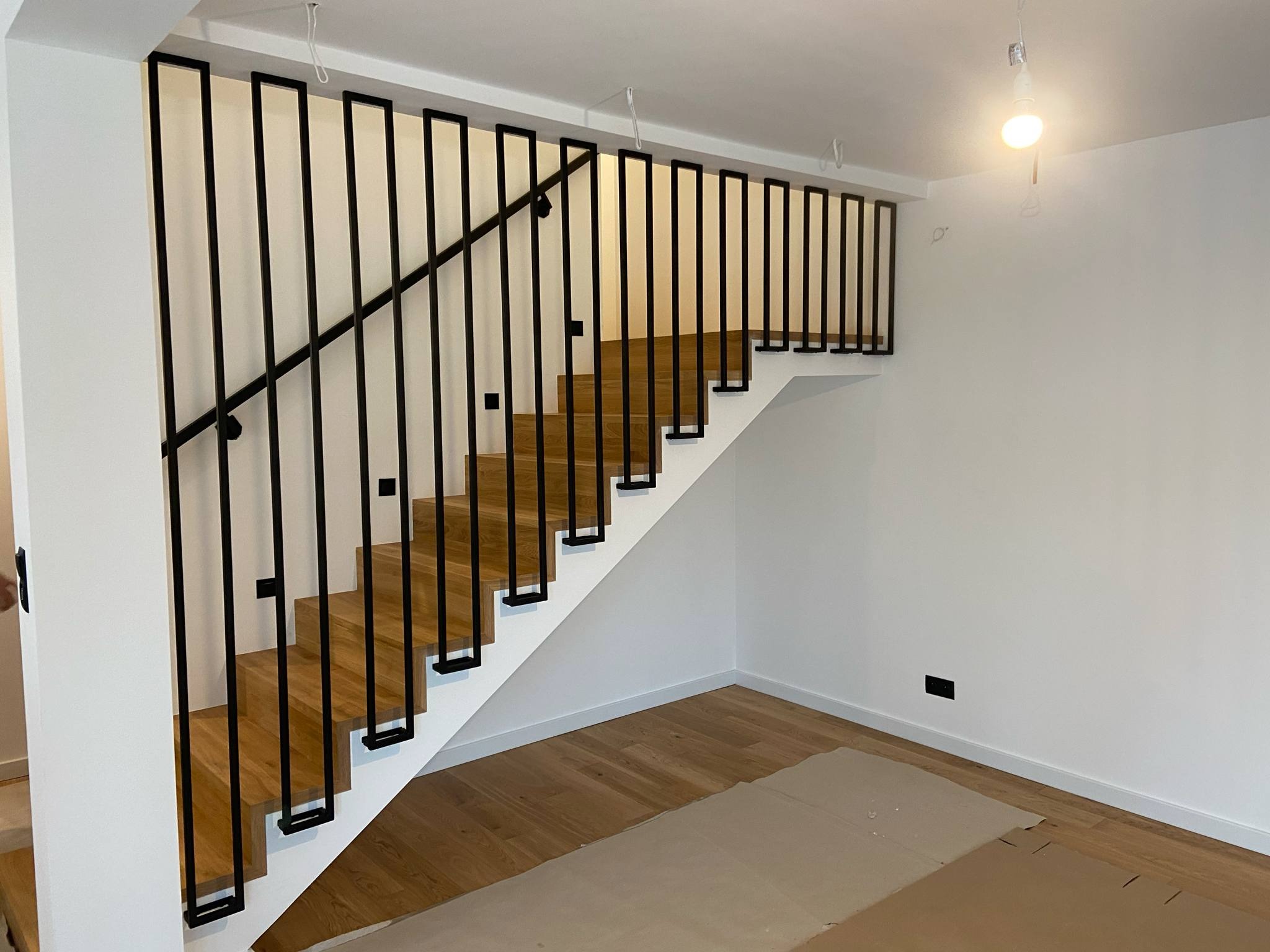 Stylowe schody z drewnianymi stopniami i czarną, minimalistyczną balustradą. Jasne wnętrze, proste linie, nowoczesny design. Częściowo zabezpieczona podłoga.