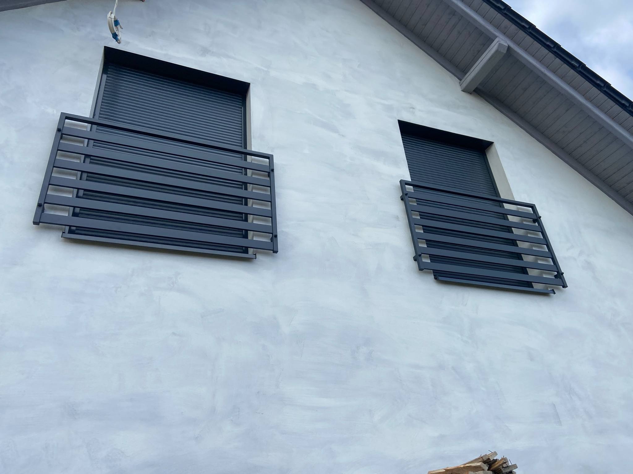 Elewacja domu z dwoma oknami z szarymi roletami i metalowymi balustradami balkonowymi. Widok z dołu, podkreślający geometrię budynku.