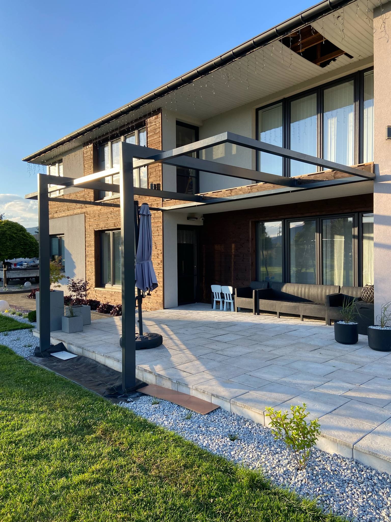 Nowoczesna pergola metalowa, zadaszenie tarasu przy domu jednorodzinnym, z meblami ogrodowymi i roślinami w donicach. Widoczne elementy wykończenia elewacji.