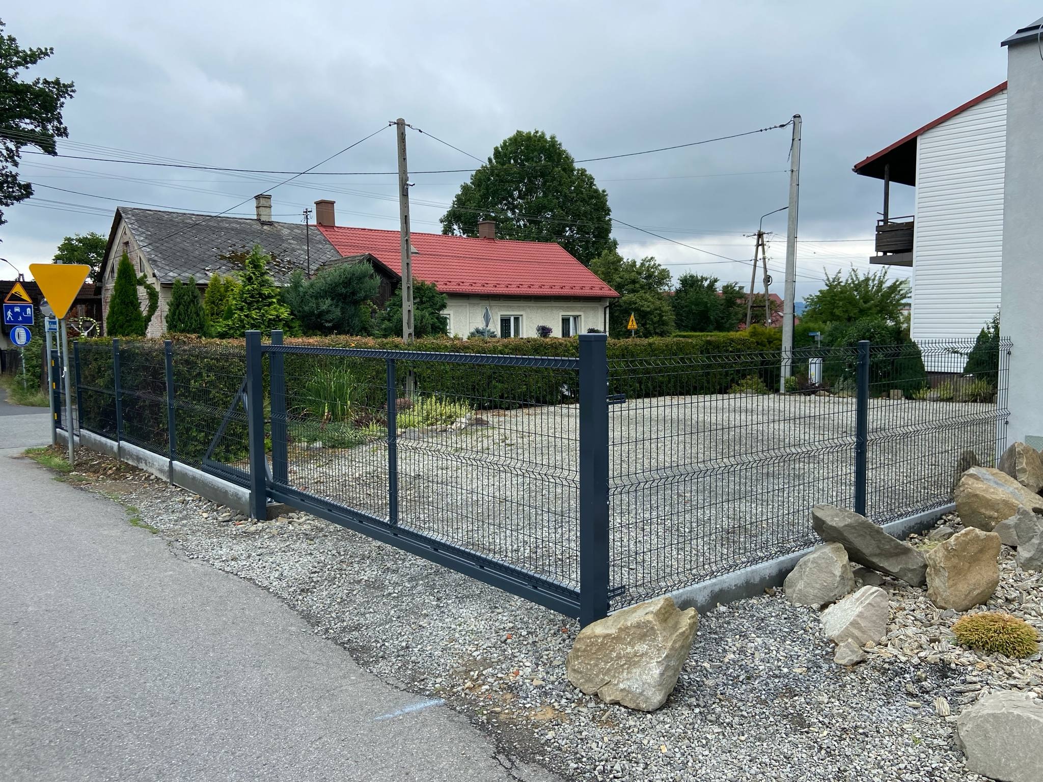 Nowoczesna, przesuwna brama panelowa w kolorze grafitowym, z niskim betonowym fundamentem i kamieniami dekoracyjnymi, na tle zabudowy jednorodzinnej.