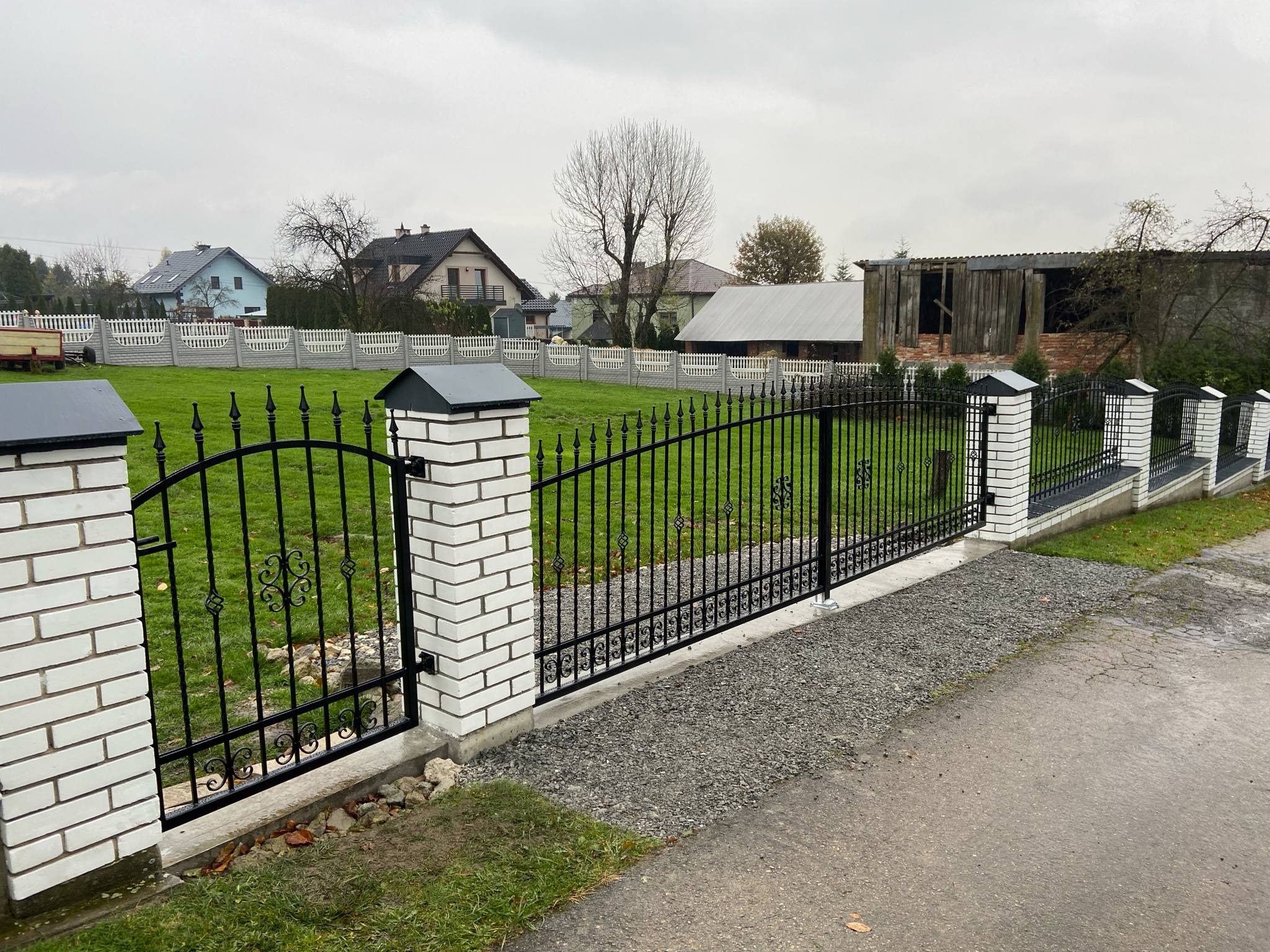 Eleganckie, czarne ogrodzenie metalowe z białymi, ceglanymi słupkami, zdobione ornamentami, na tle zielonego trawnika i zabudowań mieszkalnych w pochmurny dzień.