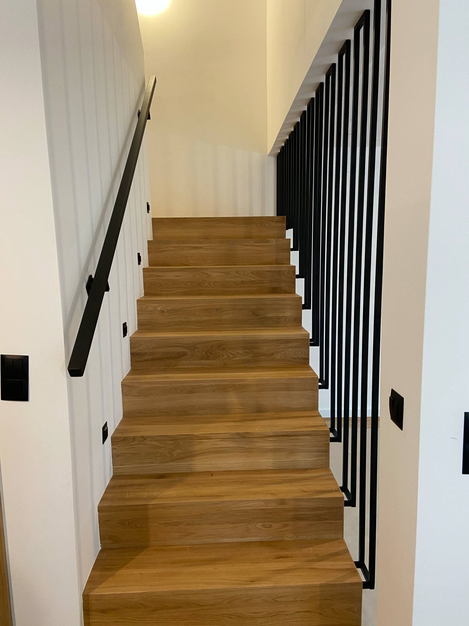 Drewniane schody w nowoczesnym wnętrzu z czarnymi, pionowymi balustradami i minimalistyczną poręczą. Oświetlenie punktowe na ścianie, jasne kolory ścian.