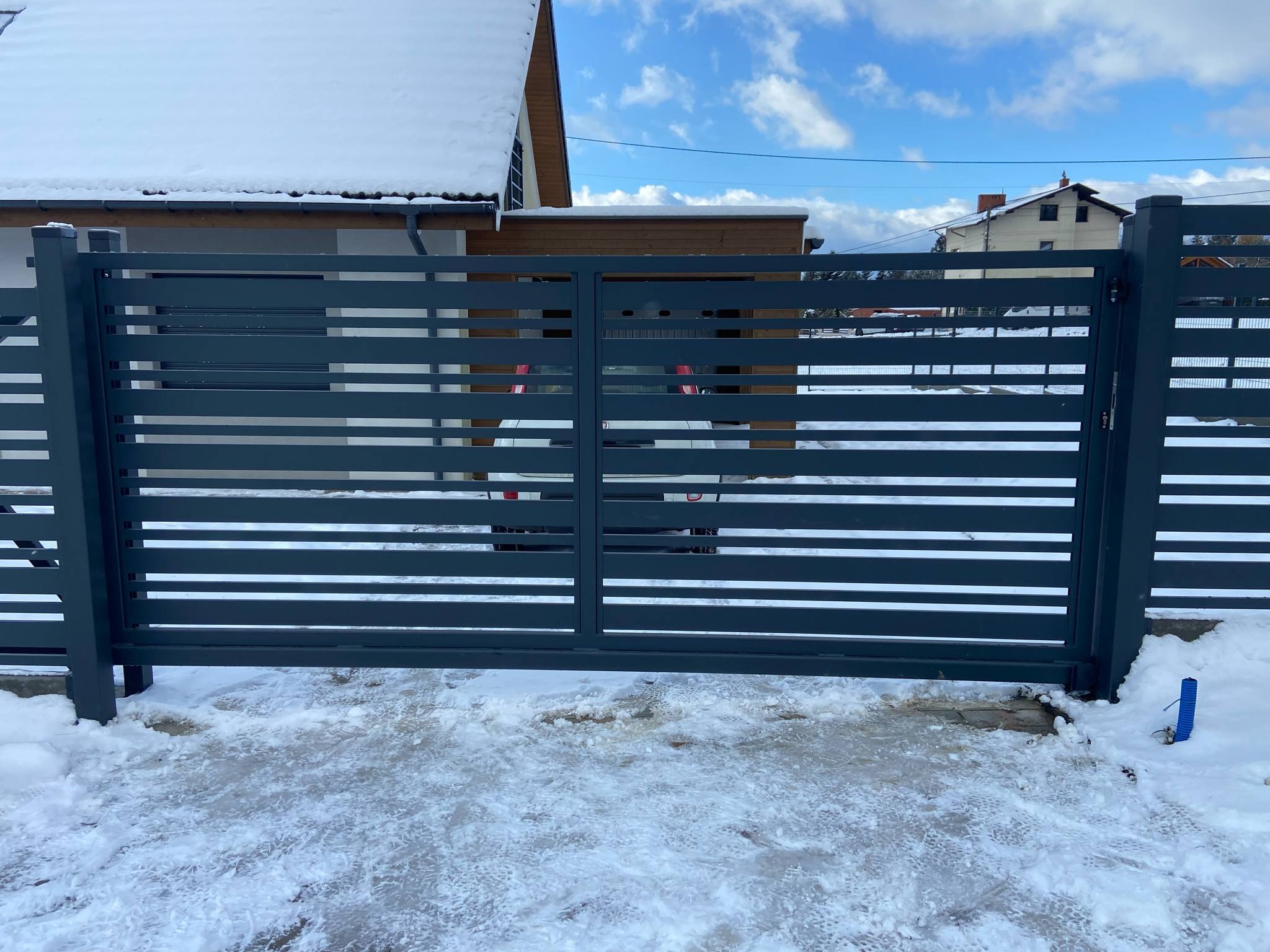 Nowoczesna, przesuwna brama wjazdowa z poziomych, szarych profili, zamontowana na posesji pokrytej śniegiem w słoneczny dzień. Widoczny dom w tle.