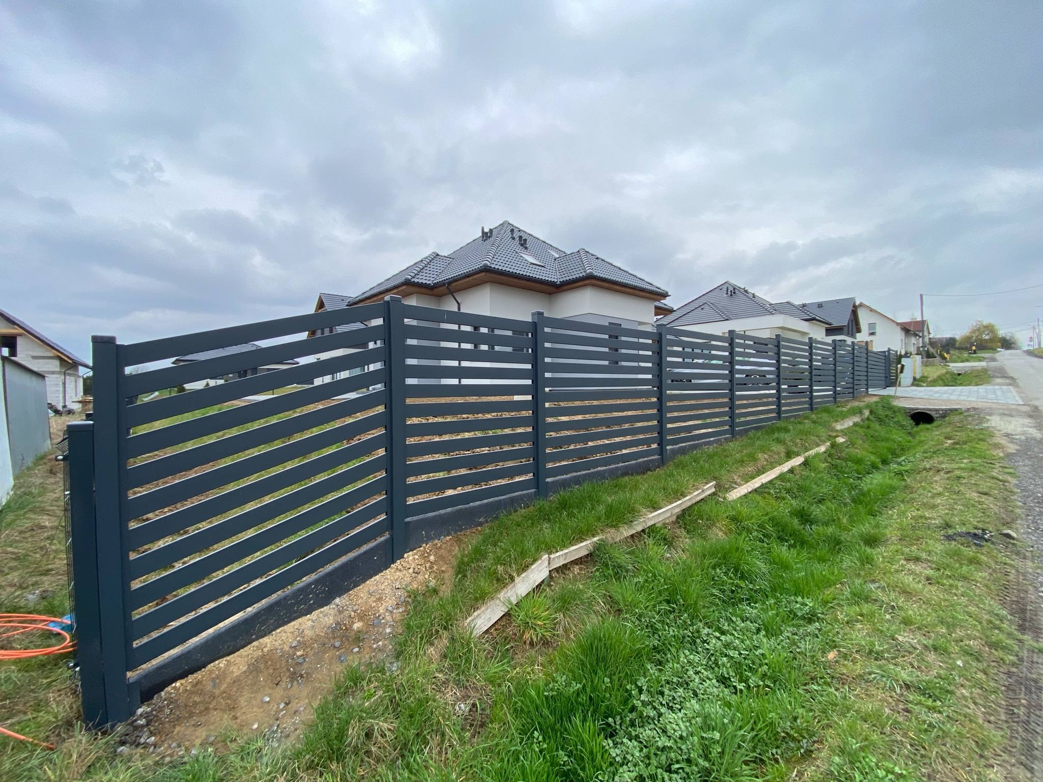 Grafitowe ogrodzenie panelowe, poziome deski, świeżo po montażu, widok z zewnątrz posesji, trawa, domy w tle, pochmurne niebo.