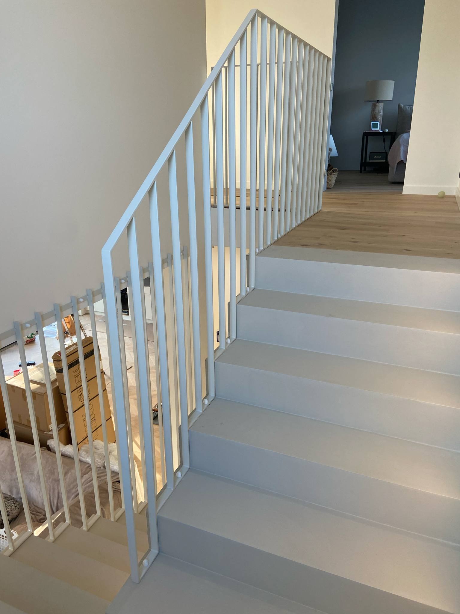 Wnętrze domu: białe, minimalistyczne schody z metalową balustradą z pionowych prętów. W tle widoczne kartony i fragment sypialni.