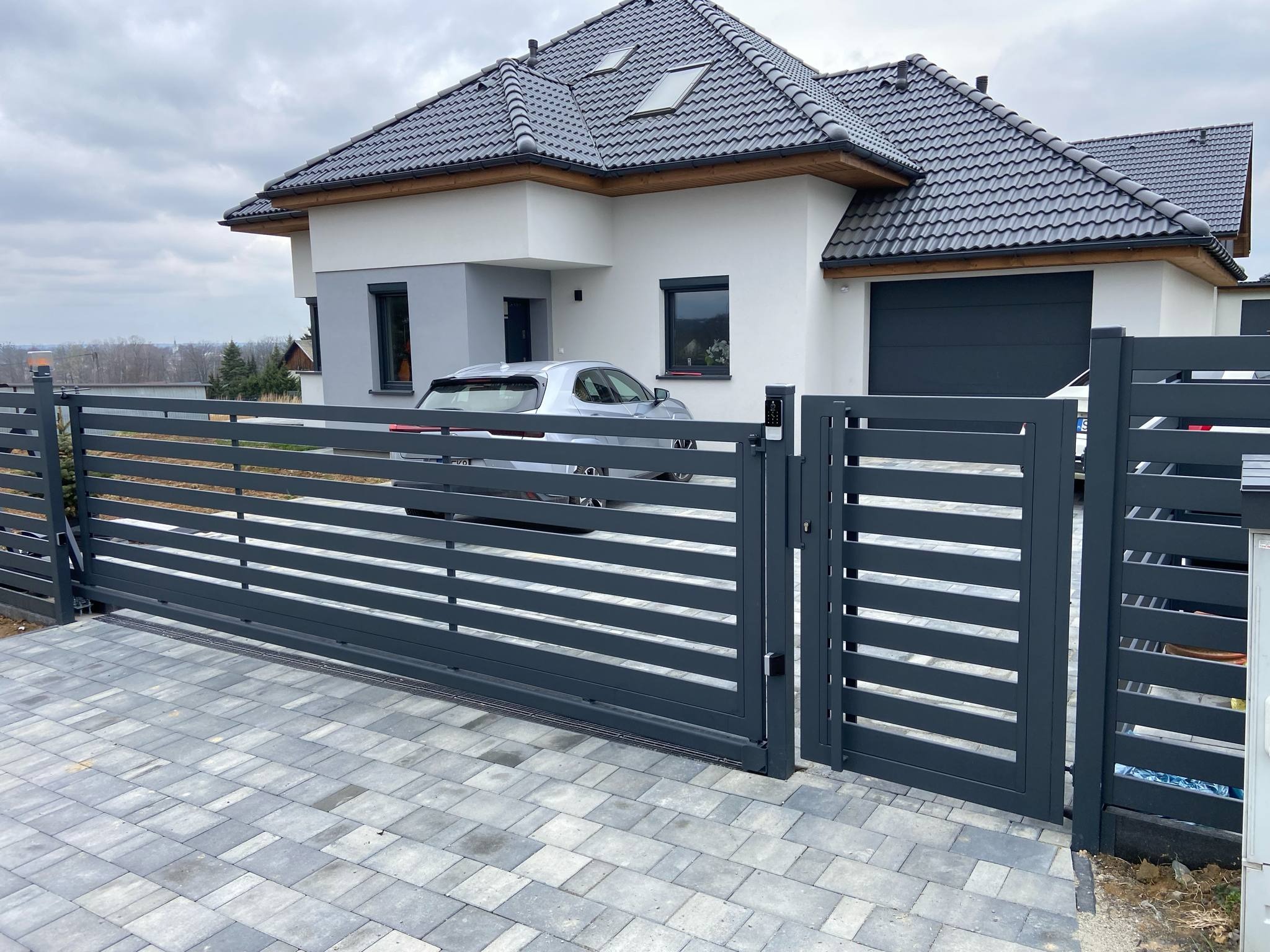 Nowoczesna, przesuwna brama wjazdowa w kolorze grafitowym z furtką, na tle domu z szarą dachówką i kostki brukowej.