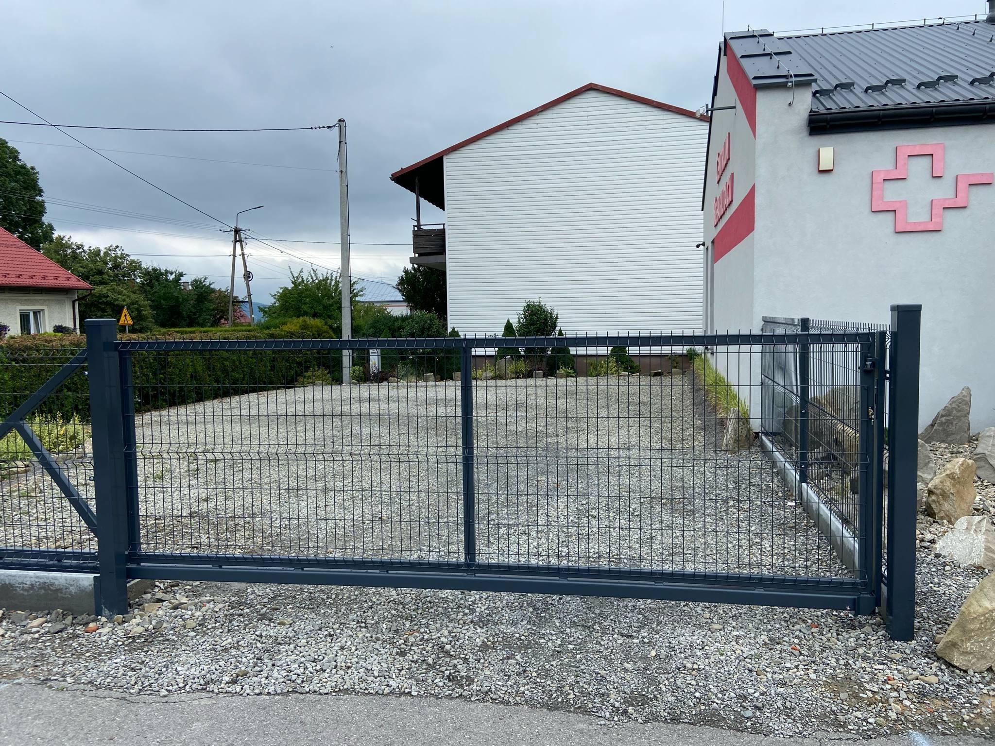 Przesuwna brama panelowa w kolorze grafitowym, z widoczną siatką i elementami mocującymi, na tle budynku z elementami czerwonymi i szarym niebem.